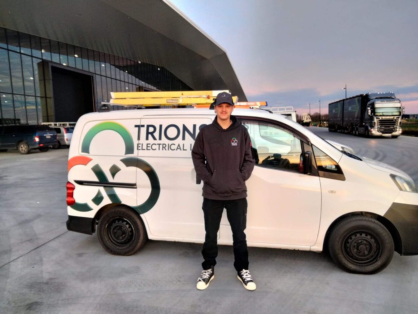 Trions technician standing in front of a branded van parked at a commercial facility, with trucks and modern buildings nearby.